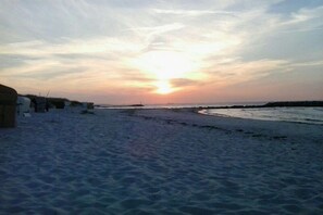 Ein Strand mit Sonnenuntergang im Hintergrund, sandigem Gelände und einem Steg, der sich ins Wasser erstreckt.