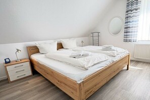 Ein ordentlich bezogenes Bett mit weißen Leinentüchern, ein hölzernes Kopfteil, ein Nachttisch mit Lampe und ein Fenster mit Vorhängen.