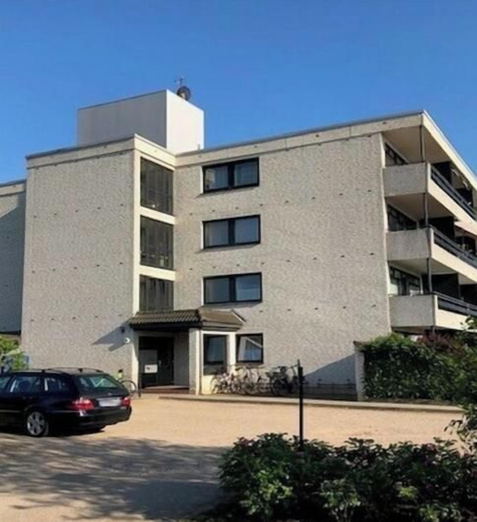 Un immeuble à plusieurs étages doté d’une façade plate, une voiture garée devant et un ciel dégagé.