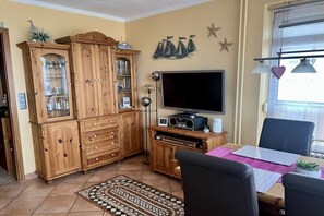 Ein Wohnzimmer mit einer Holzkassette, einem Flachbildfernseher, einem Esstisch mit Stühlen und einem gemusterten Teppich.