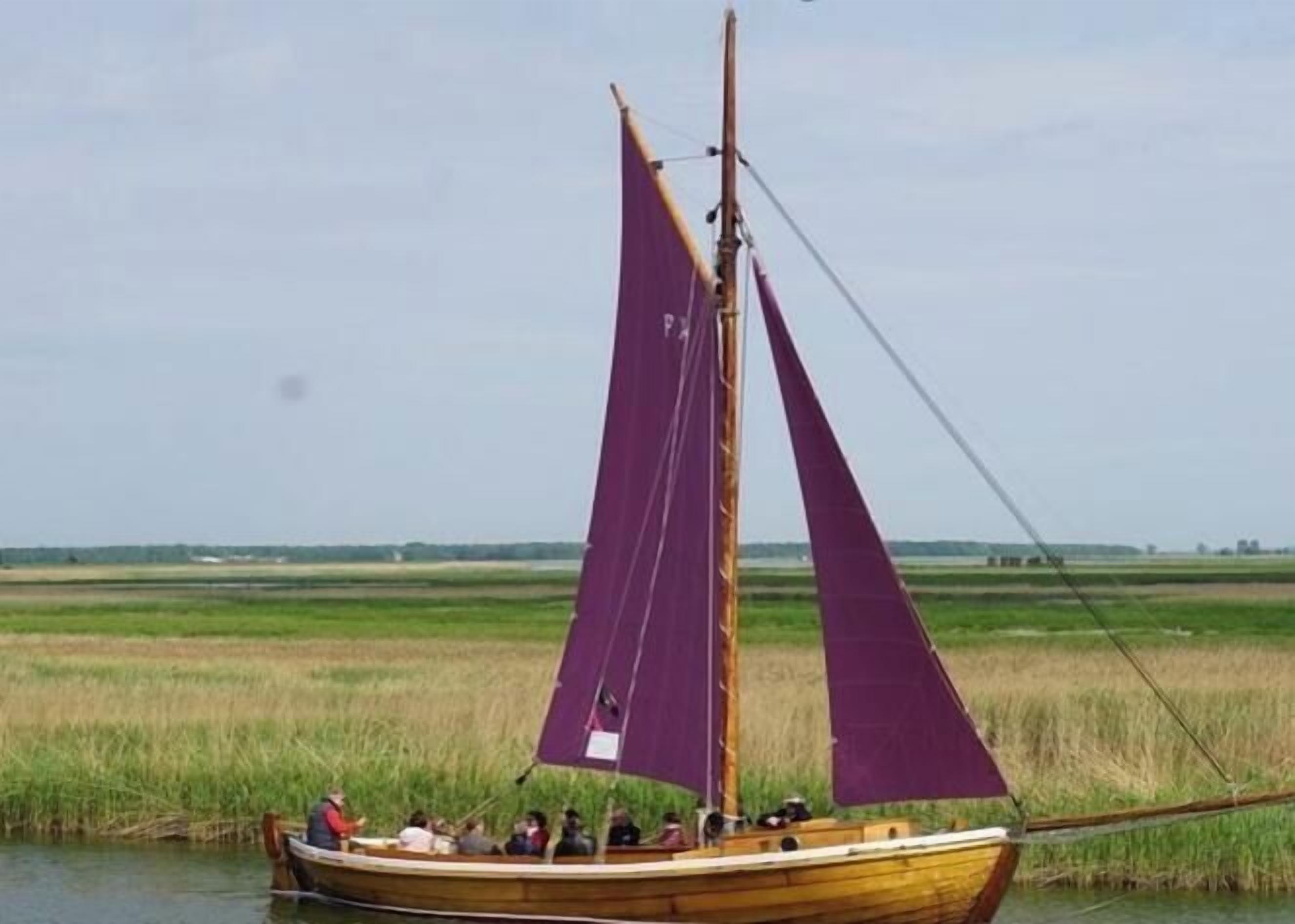 Un bateau en bois traditionnel, doté de voiles violettes, glisse sur une étendue d’eau calme, entourée de vastes champs ouverts.