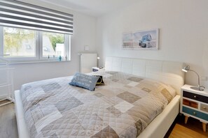 A bedroom with a bed, bedside table, lamp, and a window with blinds. - Holiday apartment with 2 bedrooms (Breege\/Juliusruh OT Juliusruh)
