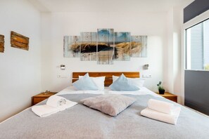 Ein Schlafzimmer mit einem großen Bett, einem Kopfteil, zwei Nachttischen, einem Wandbild und einem Fenster mit Jalousien.