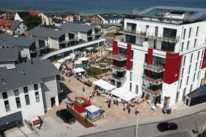 Un bâtiment moderne à plusieurs étages, un espace extérieur couvert où se trouvent des personnes et des tentes, et une vue sur la mer en arrière-plan.