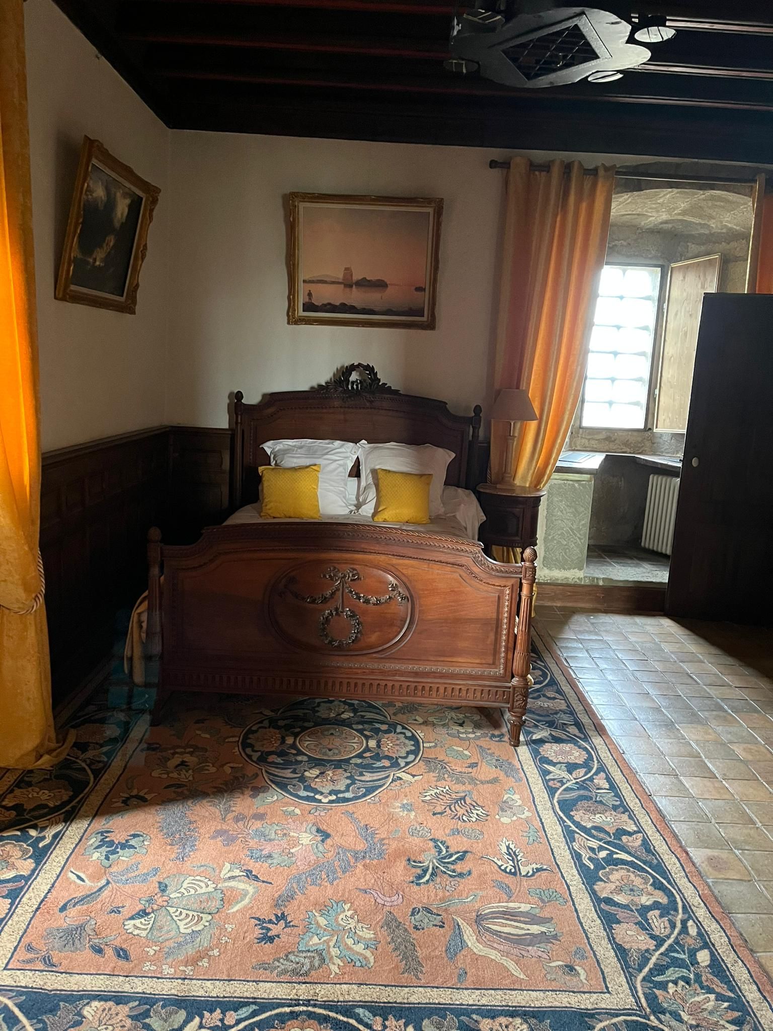 Een slaapkamer met een houten bed, een groot raam met gordijnen, een schilderij aan de muur en een kamerkleed met dessin.