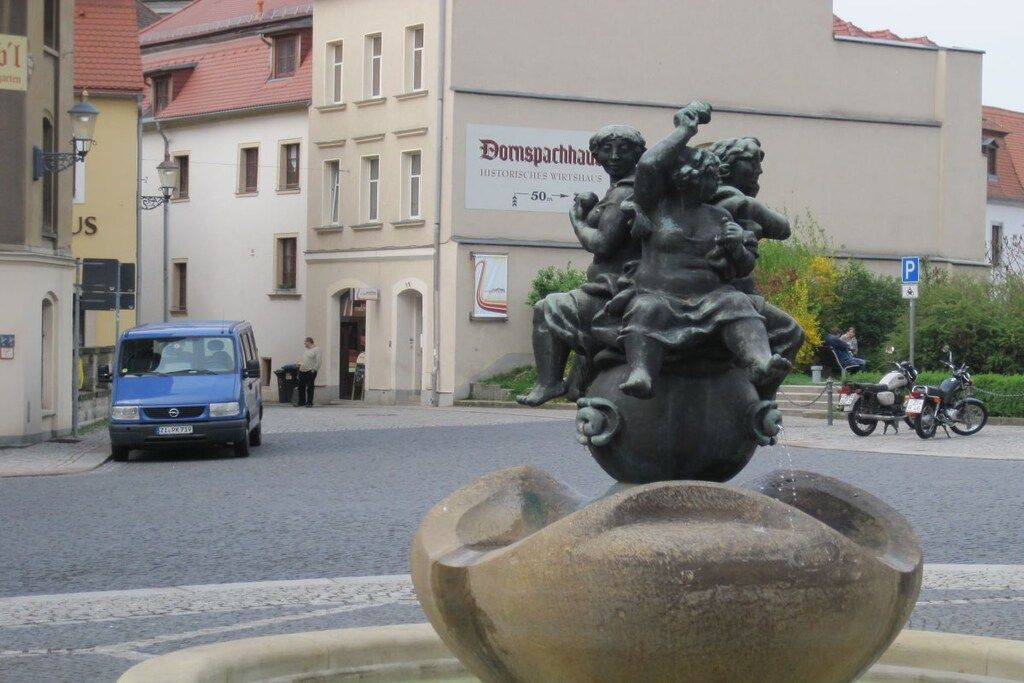 Une statue représentant deux personnages, placée sur une fontaine dans un environnement urbain, avec des bâtiments en arrière-plan.