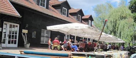 A riverside restaurant with outdoor seating, a boat docked nearby, and a building with a dark facade.