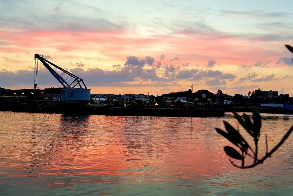A waterfront scene with a crane and buildings in the background during sunset.