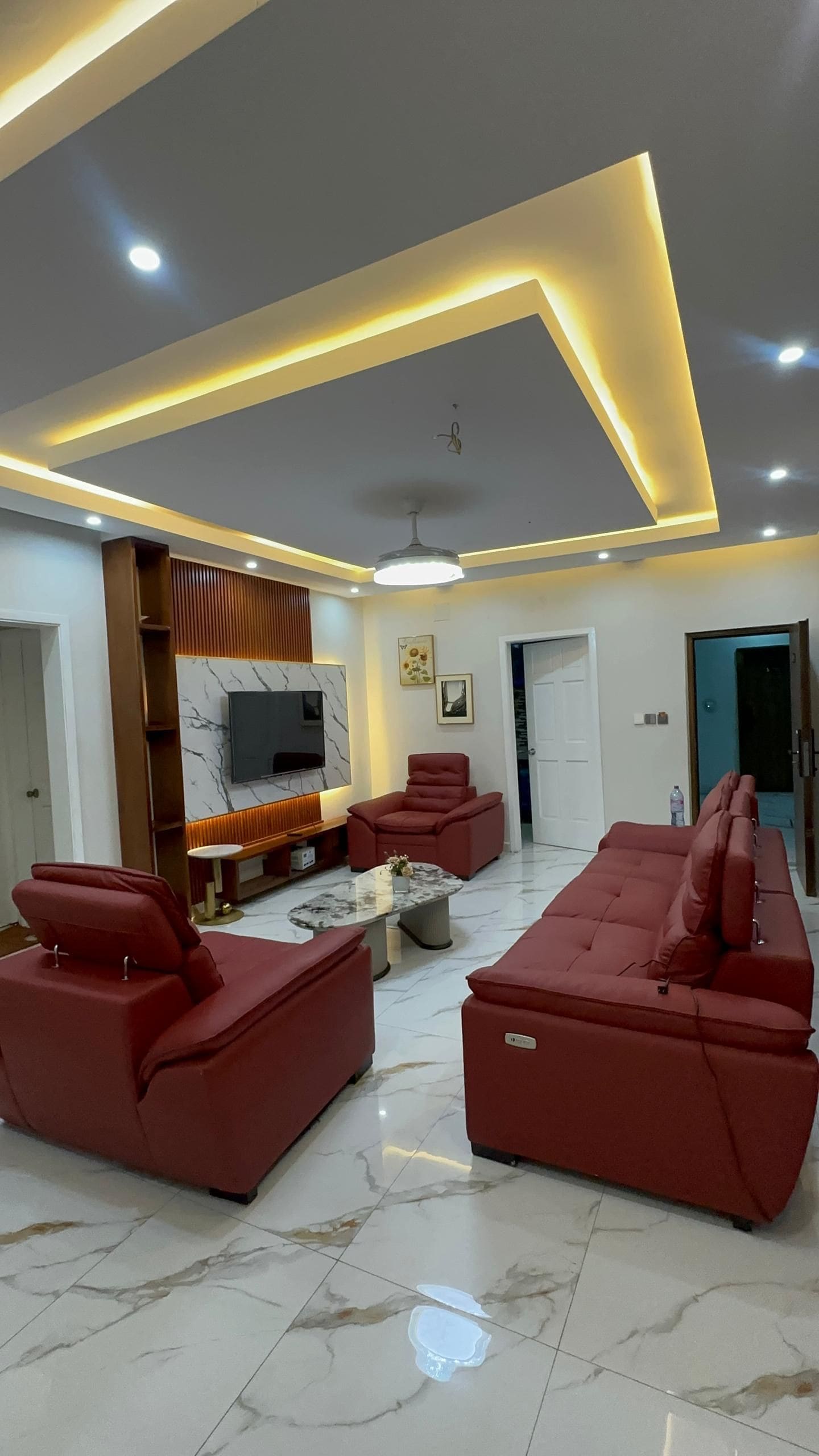 A modern living room with a sectional sofa, red armchairs, a marble coffee table, and a flat-screen TV mounted on a wall with a marble pattern.
