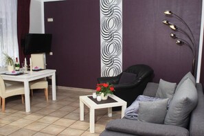 Ein Wohnzimmer mit einer Couch, einem Couchtisch mit Blumen, einem Esstisch mit Stühlen und einem Fernseher an der Wand.