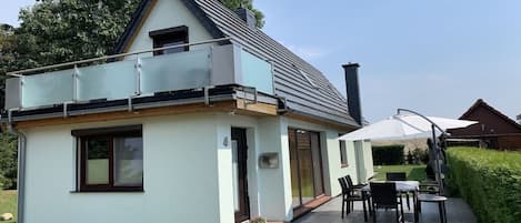 Ein zweigeschossiges Haus mit einem Balkon, einer Terrasse und Gartenmöbeln sowie einem Schornstein.