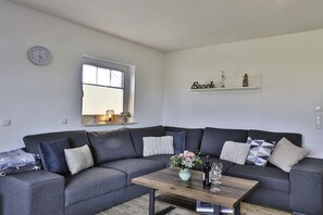 A living room with a grey sectional sofa, a wooden coffee table with a vase and wine glasses, a clock on the wall, and a shelf with decorative items. - Holiday home Backbord (Ostseebad Nienhagen)
