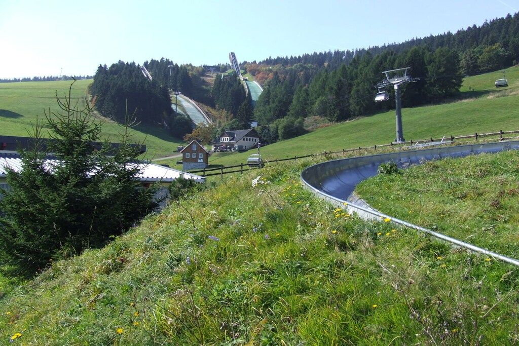 Ländliche Landschaft mit einem Haus, einem kleinen Teich und einer Seilbahn im Hintergrund.