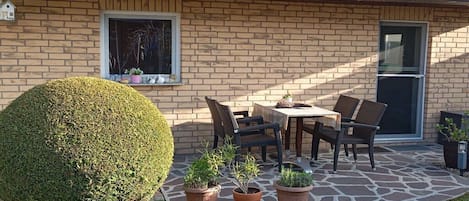 A brick house with a patio, a round hedge, and a table with chairs.