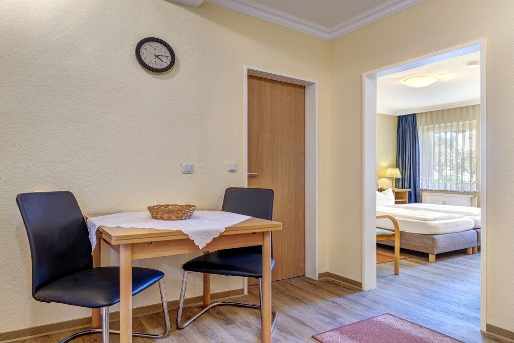 A room with a wooden table, two chairs, a clock on the wall, and a bed with a window view.