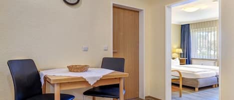 A room with a wooden table, two chairs, a clock on the wall, and a bed with a window view.