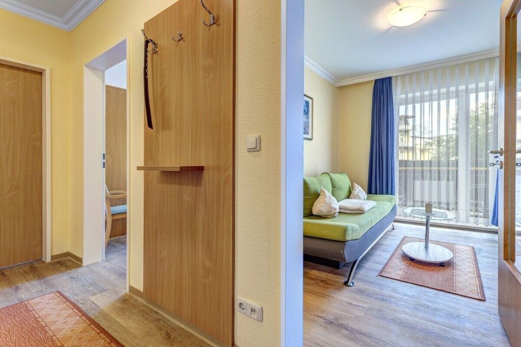 Een hotelkamer met een houten garderobe, een groene bank, een klein rond tafeltje en uitzicht buiten door grote ramen.