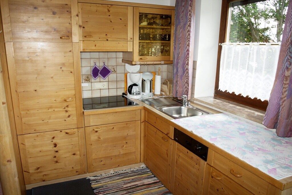 Een compacte keuken met houten kastjes, een inbouwvaatwasser en een raam met kantgordijnen.