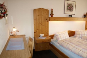 Ein kleiner, gemütlicher Raum mit einem Holztisch, einem Bett mit kariertem Bettzeug, einem Nachttisch und einem an der Wand befestigten Regal mit einer Topfpflanze.