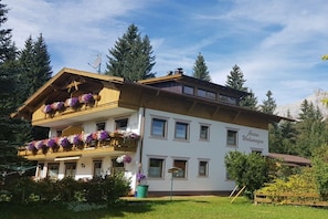 Ein zweistöckiges Gebäude mit einem an Blumen bepflanzten Balkon, einem Schild, das es als Pension kennzeichnet, und blauem Himmel.