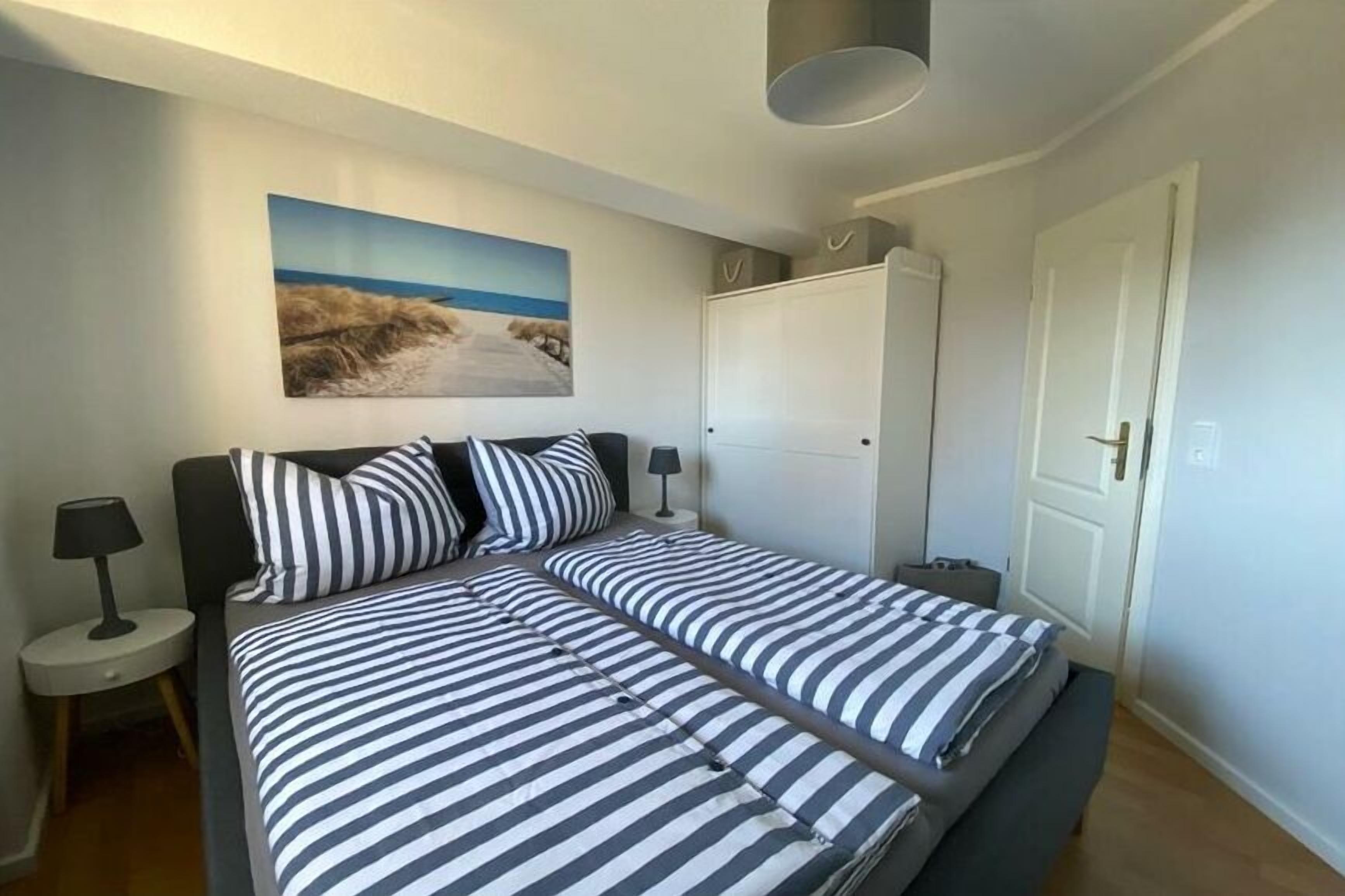 Ein Doppelbett mit gestreifter Bettwäsche, ein Nachttisch mit Lampe, ein Kleiderschrank und ein Gemälde einer Strandansicht.