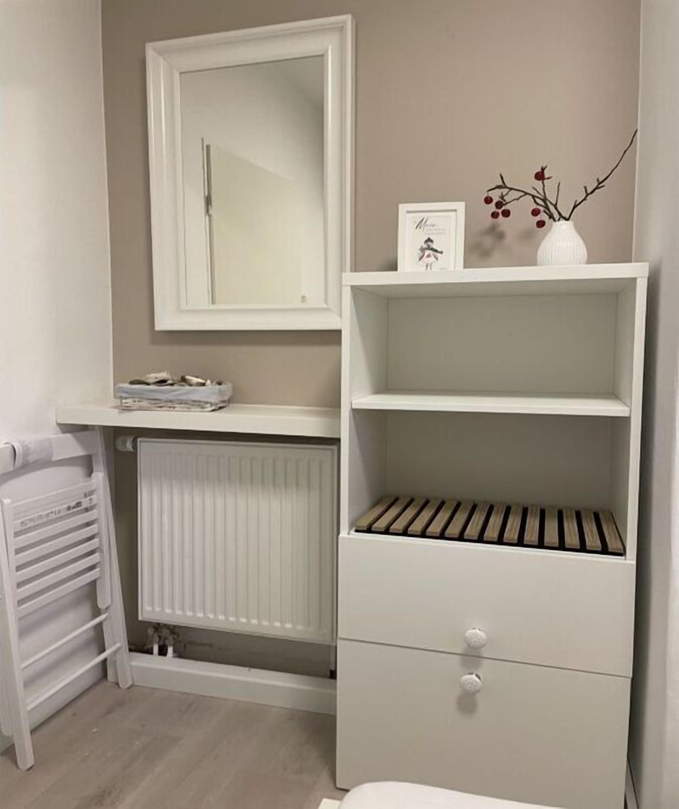 Et badeværelse med en hvid radiator, et håndklædetørrestativ, et spejl og en hylde med en billedramme og en vase.