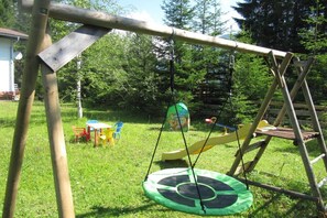 Ein Spielplatz aus Holz mit einer Schaukel, einer Rutsche und einem Tisch mit Stühlen auf einer Rasenfläche.