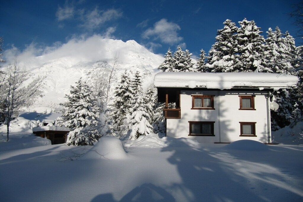 A snow-covered building with a balcony, surrounded by snow-laden trees and a mountain in the background.