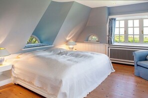 Een slaapkamer met een schuine kap, een bed met een witte dekbedovertrek, een raam met gordijnen, en een radiator.
