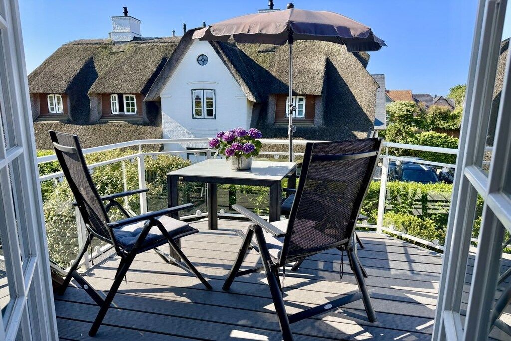 Eine Terrasse mit zwei Stühlen und einem Tisch, einer Kübelpflanze und einem Haus mit Strohdach im Hintergrund.