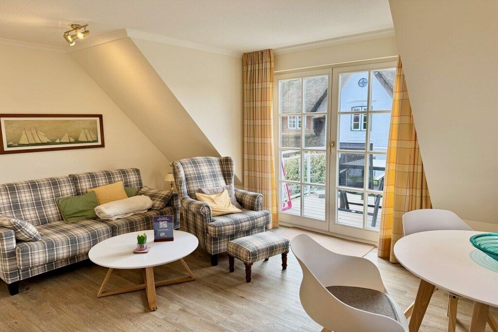 Een gezellige woonkamer met een bank, fauteuil, pouf en salontafel, een raam met gordijnen en een schilderij aan de muur.