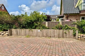 Ein gepflasterter Weg führt zu einem Holzzaun, im Hintergrund ist ein Haus zu sehen.
