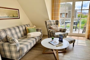 Une pièce avec un canapé à carreaux, un fauteuil assorti, une table basse ronde sur laquelle se trouvent un livre et une télécommande, et une grande fenêtre donnant sur une maison et de la verdure.