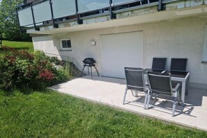 A residential building with a garage door, a small patio with a table and chairs, and a garden area with bushes and flowers. - Holiday apartment with 2 bedrooms (Altenberg)