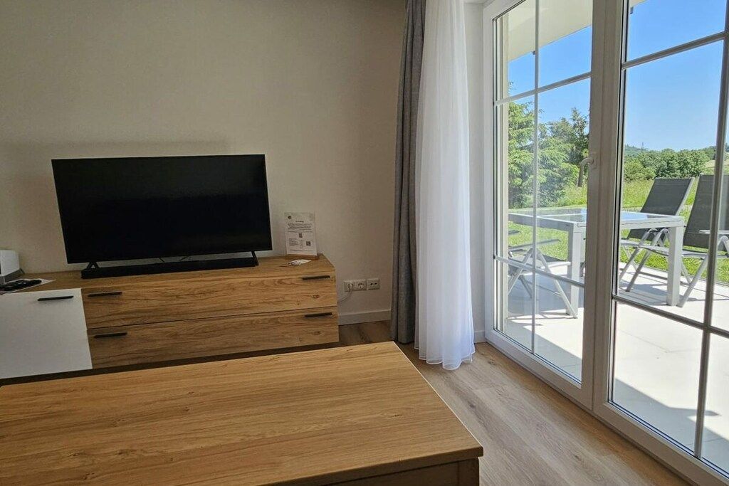 A room with a flat-screen TV on a wooden dresser, a white desk, and a large window with a view of outdoor furniture.