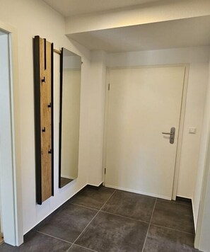 Een gang met een witte deur, een spiegel met houten lijst en donkere betegelde vloer.