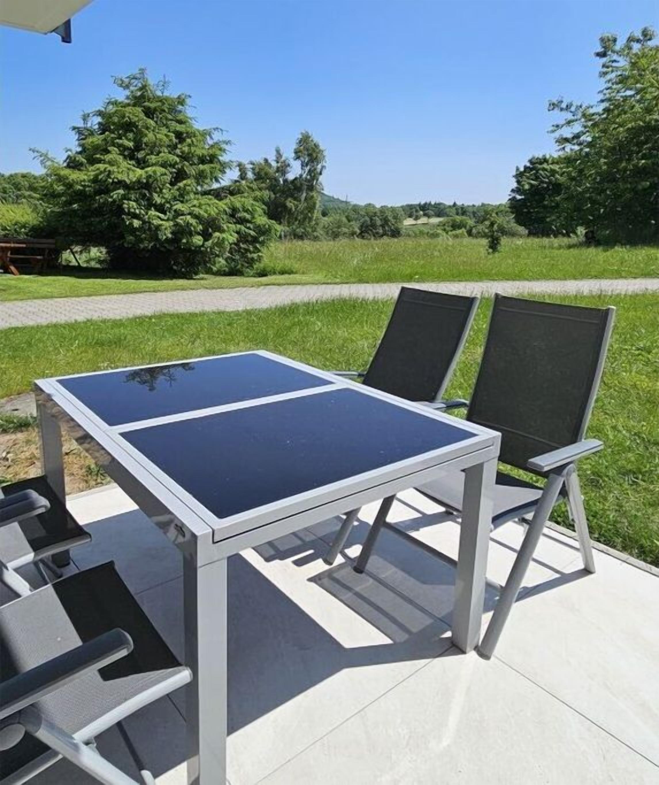 An outdoor table with chairs, a paved area, and a grassy field with trees in the background.