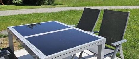 An outdoor table with chairs, a paved area, and a grassy field with trees in the background.