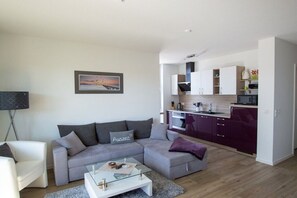 A modern living room with a grey sofa, a white coffee table, and a kitchen area with purple cabinets and white appliances. - Holiday apartment in Glowe with swimming pool (Glowe)