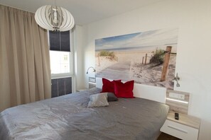 Ein Schlafzimmer mit einem großen Bett, einem Wandbild mit Strandmotiv, einem Fenster mit Vorhängen und einer modernen Pendelleuchte.