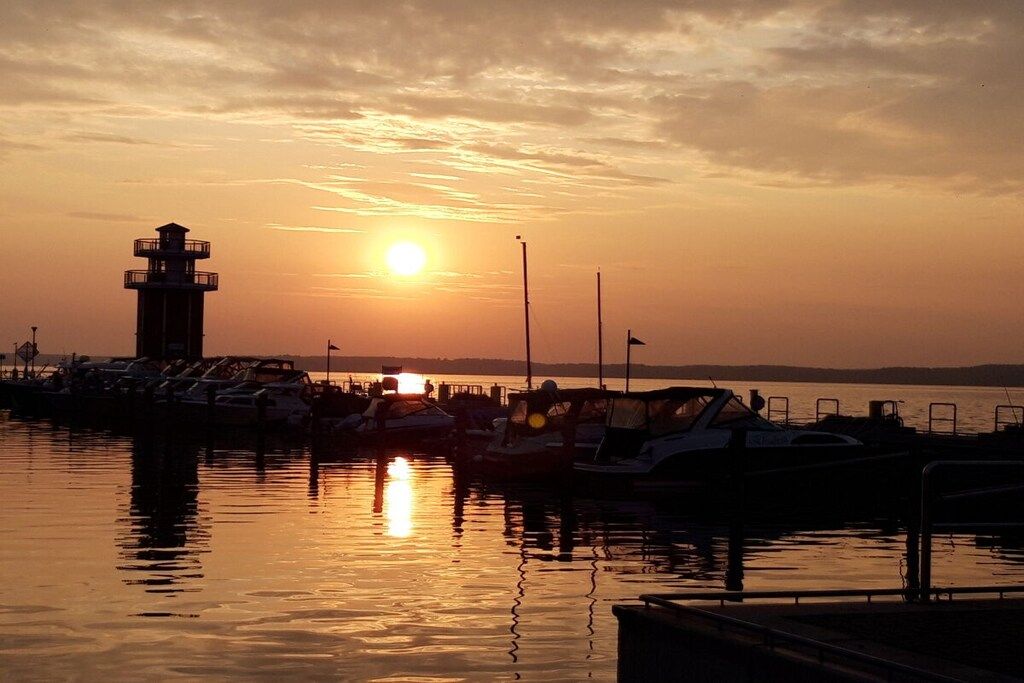 A marina at sunset with boats docked, a lighthouse, and calm water reflecting the sky.