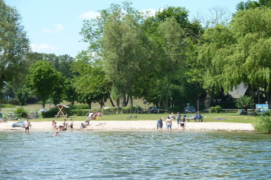 Un cadre idyllique au bord d’un lac, avec des personnes pratiquant des activités nautiques, des arbres et une zone sablonneuse équipée de chaises de plage.