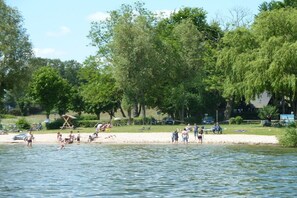 A lakeside setting with people engaging in water activities, trees, and a sandy area with beach chairs. - Holiday home with 2 bedrooms (Plau am See)