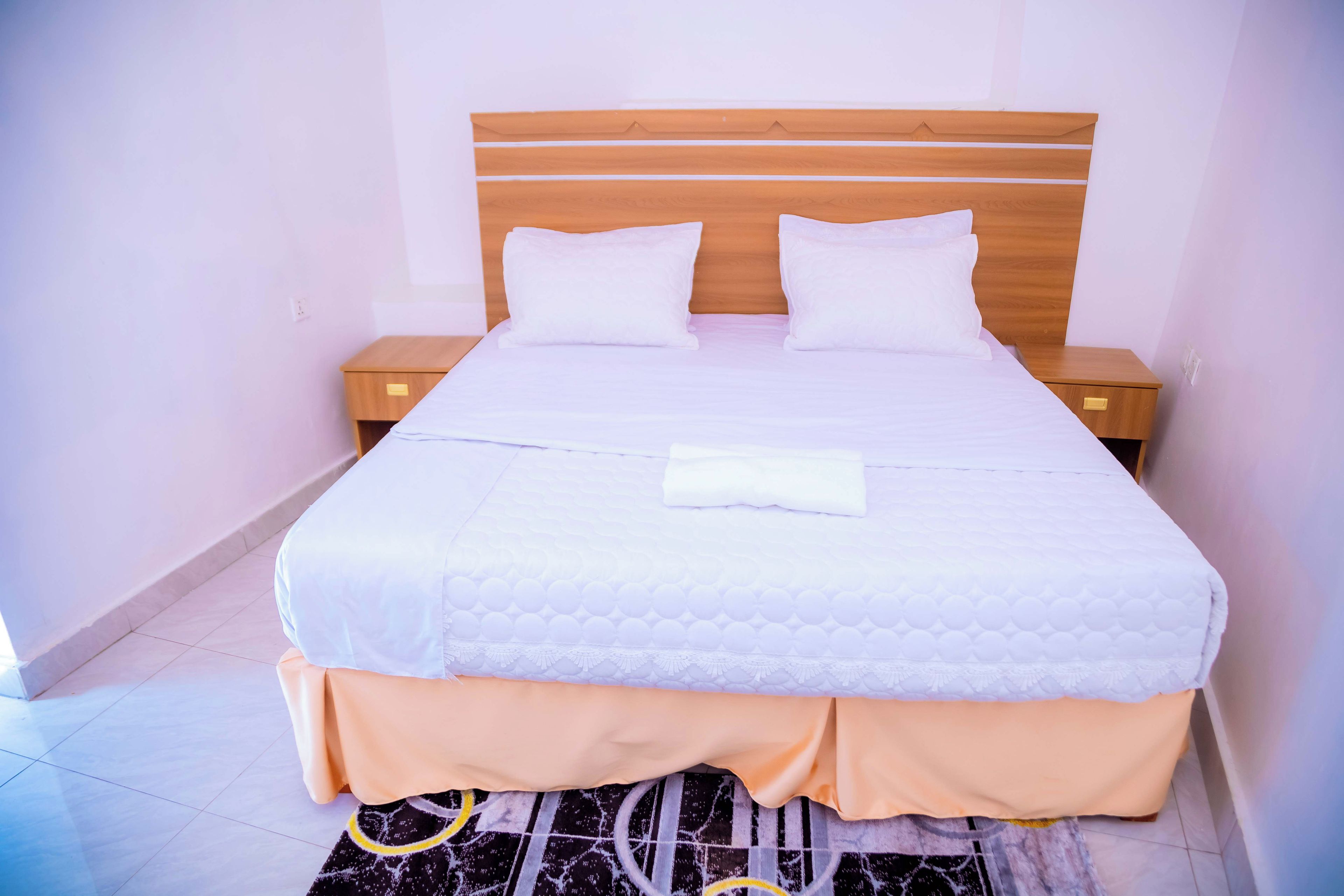 Una cama bien hecha con ropa de cama blanca y un cojín a juego, flanqueada por mesitas de noche de madera con cajones, en una habitación con paredes lisas y suelo de baldosas.