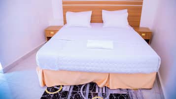 Una cama bien hecha con ropa de cama blanca y un cojín a juego, flanqueada por mesitas de noche de madera con cajones, en una habitación con paredes lisas y suelo de baldosas.