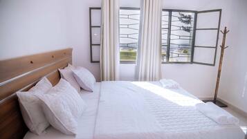 Una cama bien hecha con sábanas y almohadas blancas, un cabecero de madera y vistas a un balcón a través de una ventana con celosía metálica.