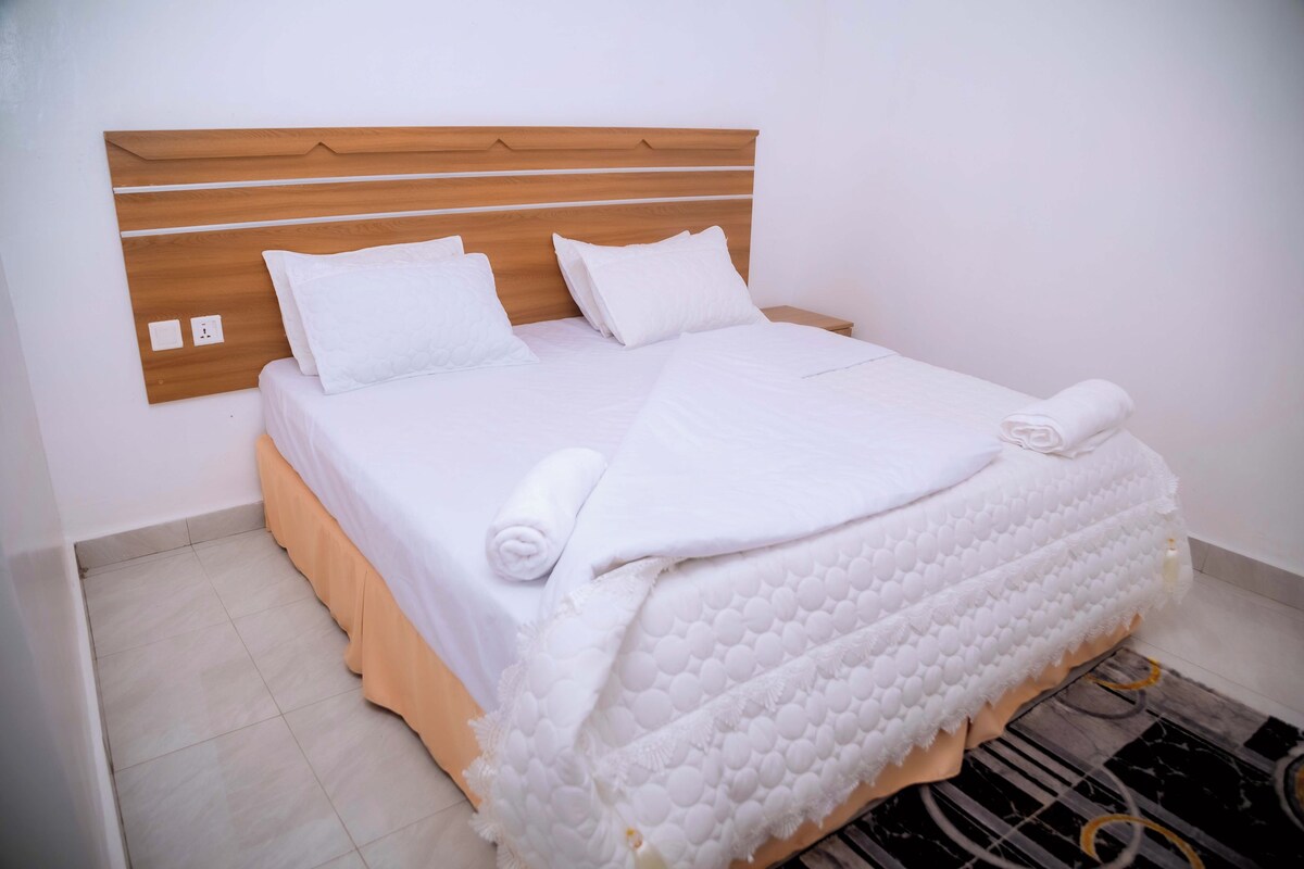 Una cama bien hecha con ropa blanca y almohadas, un cabecero de madera y una mesita de noche pequeña.