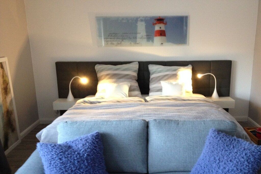 A bedroom with a bed, two bedside lamps, a wall-mounted artwork of a lighthouse, and a blue knitted throw on the bed.