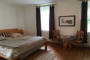 Ein Schlafzimmer mit einem Bett, zwei Stühlen, einem kleinen Tisch, einem Fenster mit Vorhängen und einem gerahmten Bild an der Wand.