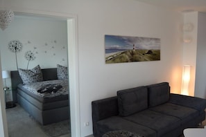 Ein modernes Wohnzimmer mit einem grauen Sofa, einem Bett mit gemusterten Betttextilien, einem Wandbild von einer Küstenlandschaft und einer Stehlampe.